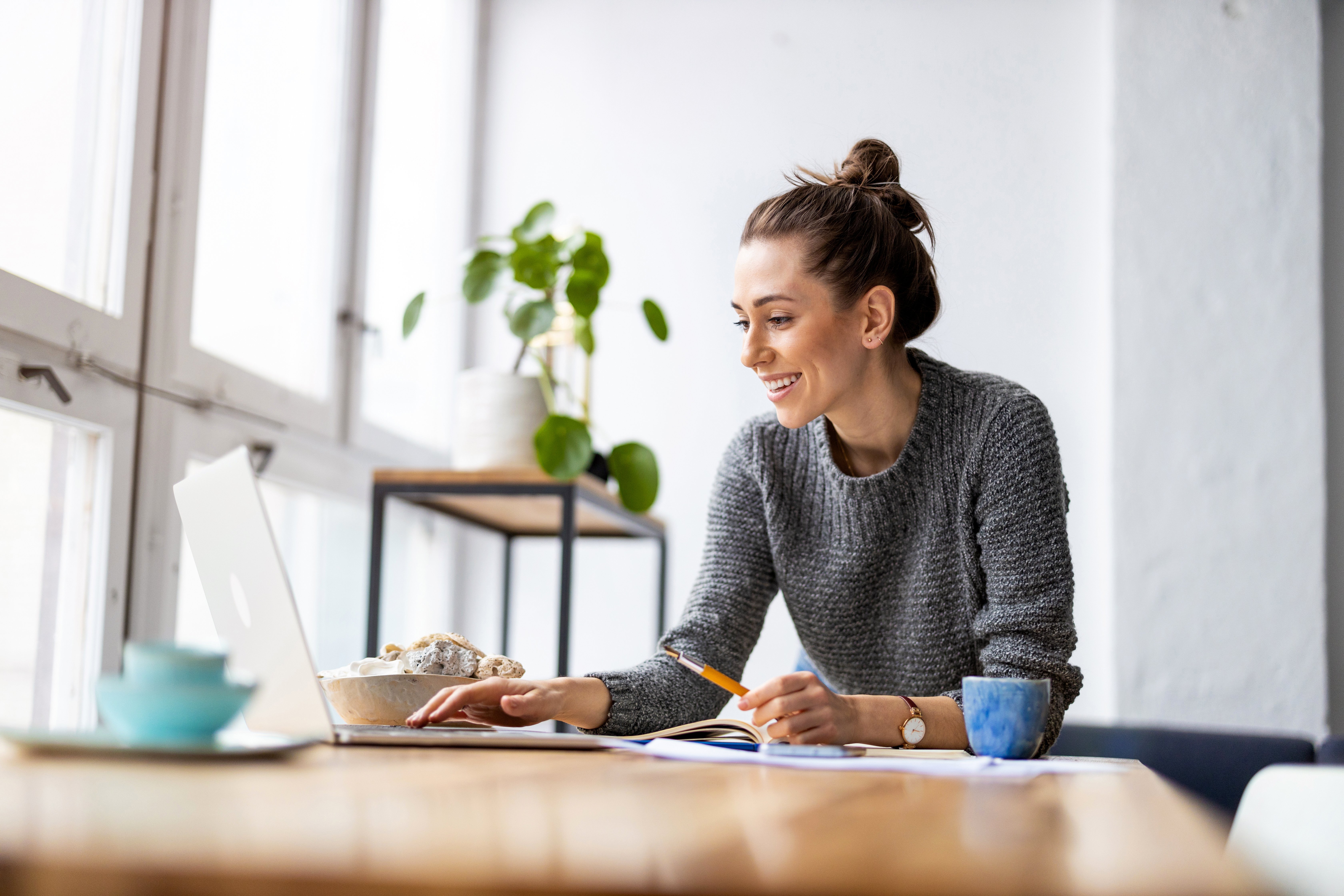 A woman with a bun hairstyle smiles while working on a laptop, sitting at a wooden table with a cup of coffee. There are plants and decorative items in the background.