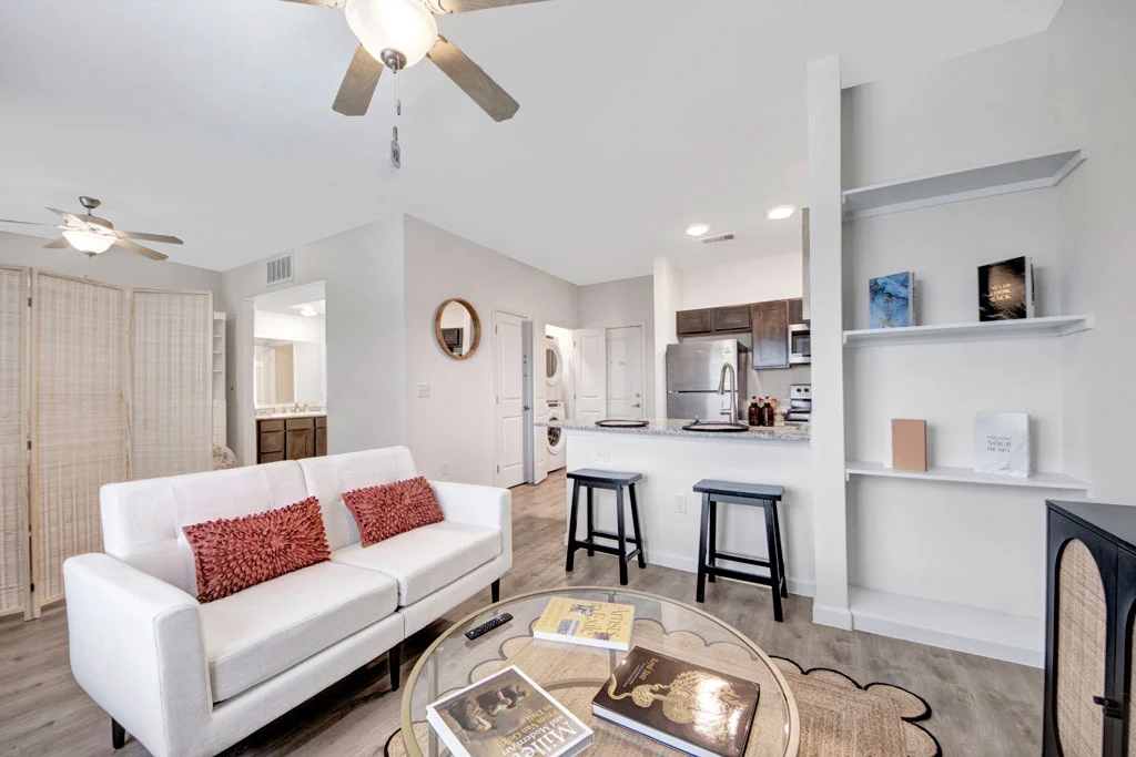 Modern Living Room Interior A bright and modern living room featuring a white sofa with red cushions, a glass coffee table with books, kitchen area in the background, and shelves displaying some decor items.
