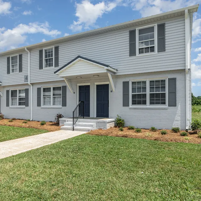 Exterior view of a townhouse with two stories, featuring light gray siding, dark blue doors, and landscaped front yard.