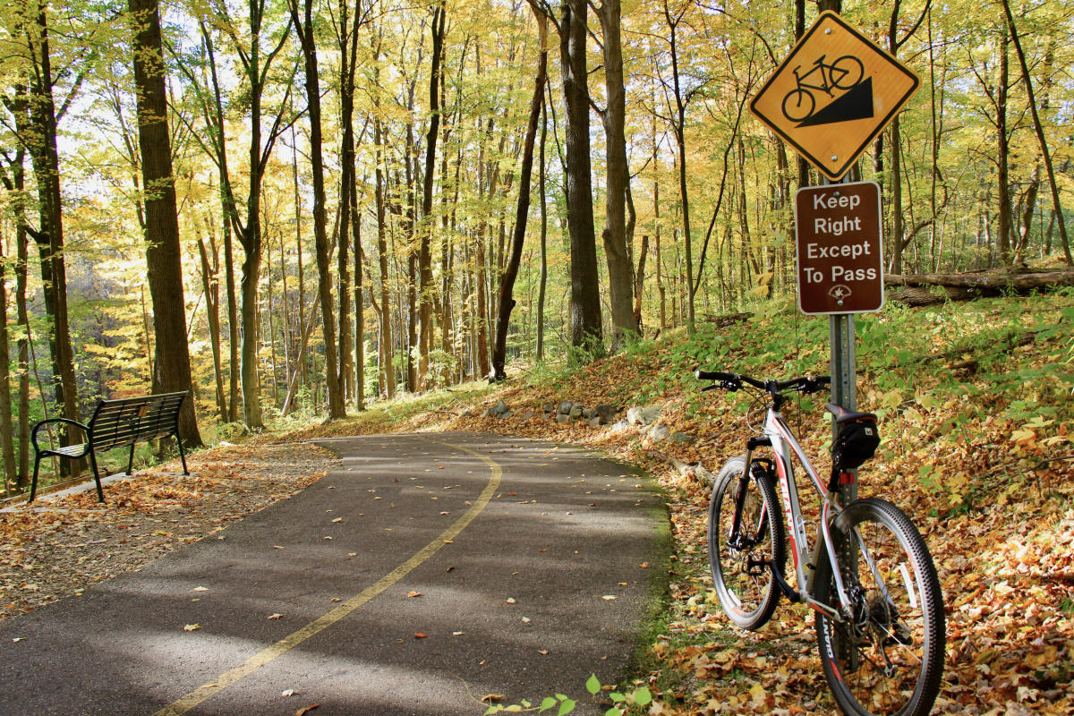 A bicycle parked beside a paved trail in a wooded area during autumn. Leaves are yellow and orange on the trees, creating a picturesque fall scene. A warning sign instructs to keep right except to pass. A bench is visible on the side of the path.