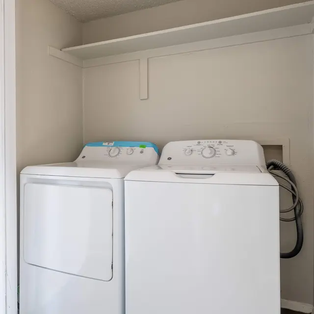 A compact laundry room with a white washer and dryer side by side, featuring a shelf above them in a light-colored space.