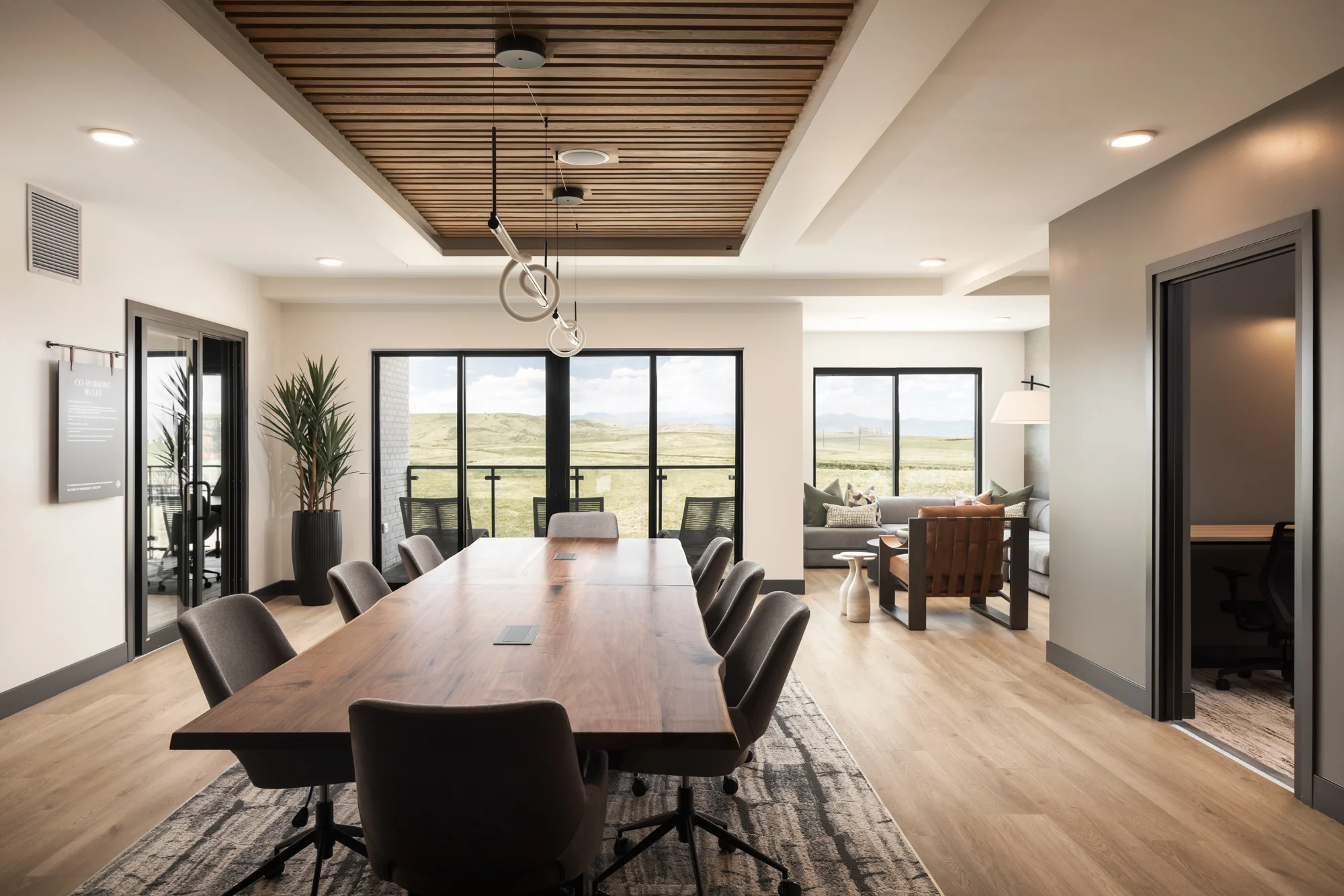 Modern Community Meeting Room A spacious community meeting room with a large wooden table surrounded by chairs. Large windows offer a view of the outdoors. There are seating areas and decorative plants in the background.