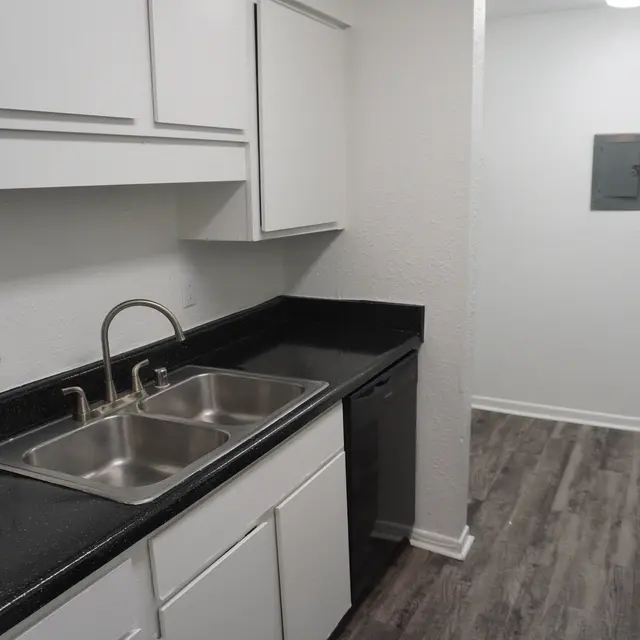 A modern kitchen featuring a double sink with chrome faucets, white cabinets, and black countertops. A black refrigerator is visible on the right, and there is an open space leading to another room.