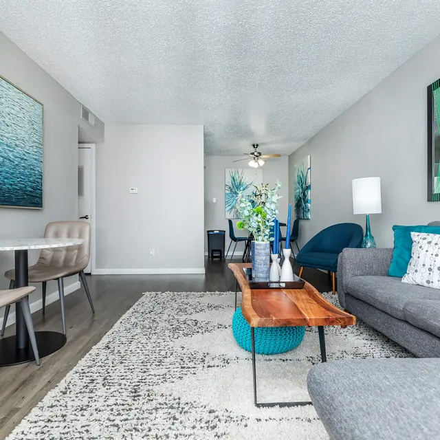 A stylish modern living room featuring a gray sofa with decorative cushions, a wooden coffee table, and an area rug. The walls are adorned with blue-toned artworks and a potted plant is visible. There is a dining area with beige chairs and a small table, and decorative elements consistent with a modern aesthetic.