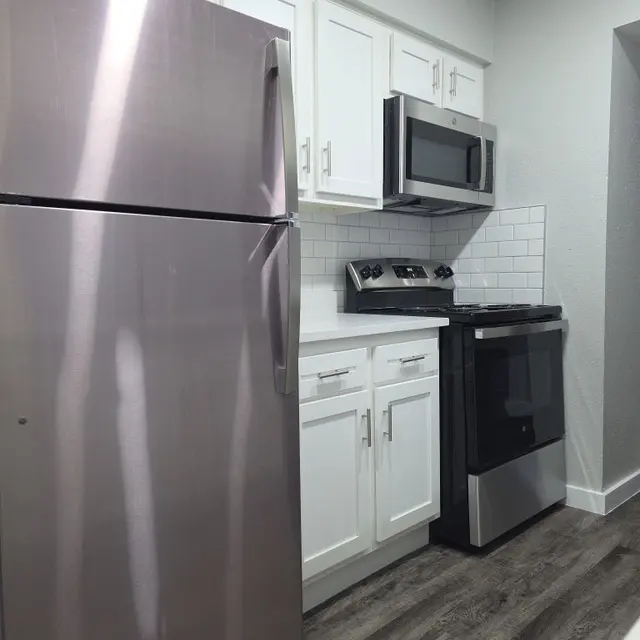 A modern kitchen featuring stainless steel appliances including a refrigerator, microwave, and stove. The cabinetry is white with a clean finish, and the floor is made of dark wood.
