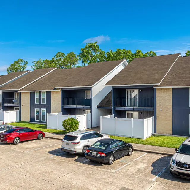 An aerial view of a multi-unit apartment complex featuring three buildings with a combination of brick and siding, surrounded by parked cars and greenery.