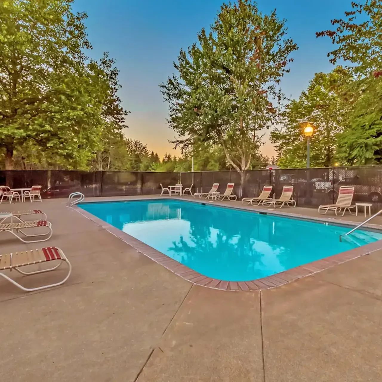 A clear swimming pool surrounded by lounge chairs and trees, with a sunset sky in the background.