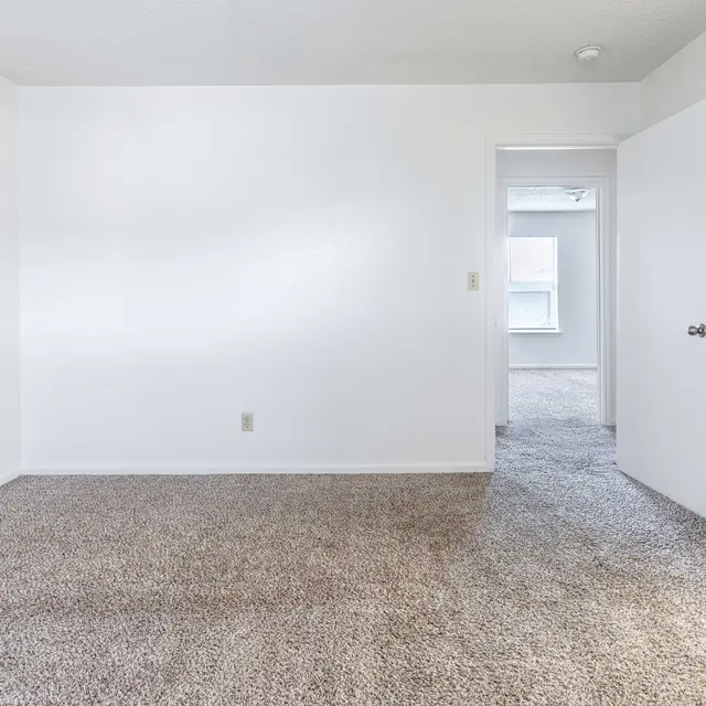 A spacious, empty room with carpet flooring and white walls. A closed door is visible on the right, leading to an adjacent room.