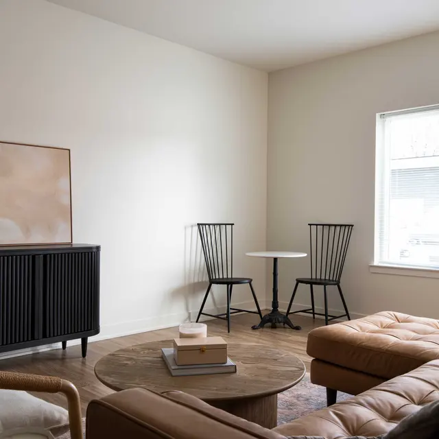 A modern and minimalist living room featuring a tan leather sofa, a round wooden coffee table, a black cabinet, two black armchairs, and a small round table by the window.