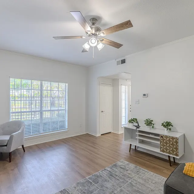 A spacious modern living room featuring a light-colored wall, a ceiling fan with wooden blades, and a large window with blinds. There is a small desk with a chair on the left side, and a white shelf unit. A gray couch is in the foreground with a yellow throw blanket.