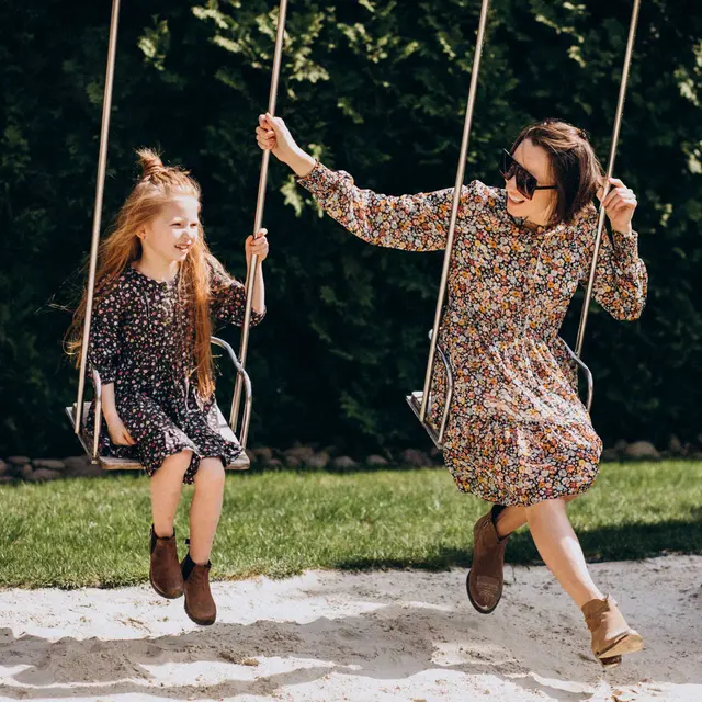 A mother and daughter enjoying a sunny day on swings in a playground. Both are wearing floral dresses and the mother is wearing sunglasses. They are seated on swings, and the backdrop features green bushes.