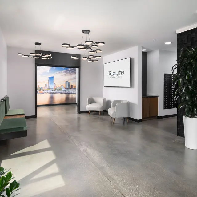 A modern office lobby featuring a sleek design with a concrete floor, contemporary furniture, and a large screen displaying a cityscape. Natural light floods the space from large windows, accented by green plants.