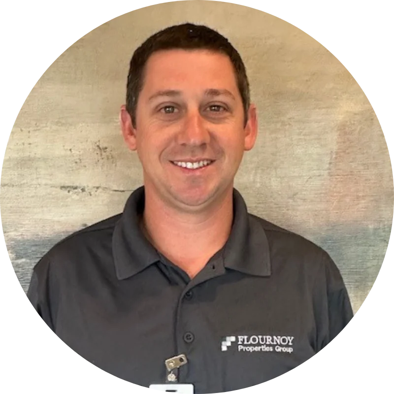 A smiling man in a dark gray polo shirt with the logo of Flournoy Properties Group. His hair is short and he stands against a textured background.
