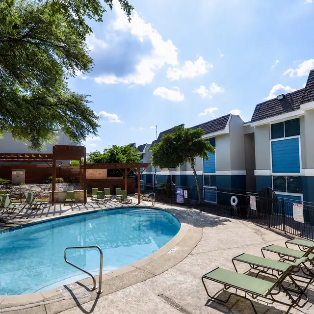 A swimming pool area featuring a rectangular pool, surrounded by green lounge chairs and an outdoor seating area beneath a wooden pergola. The background includes modern buildings with blue accents and a lush green tree overhead.