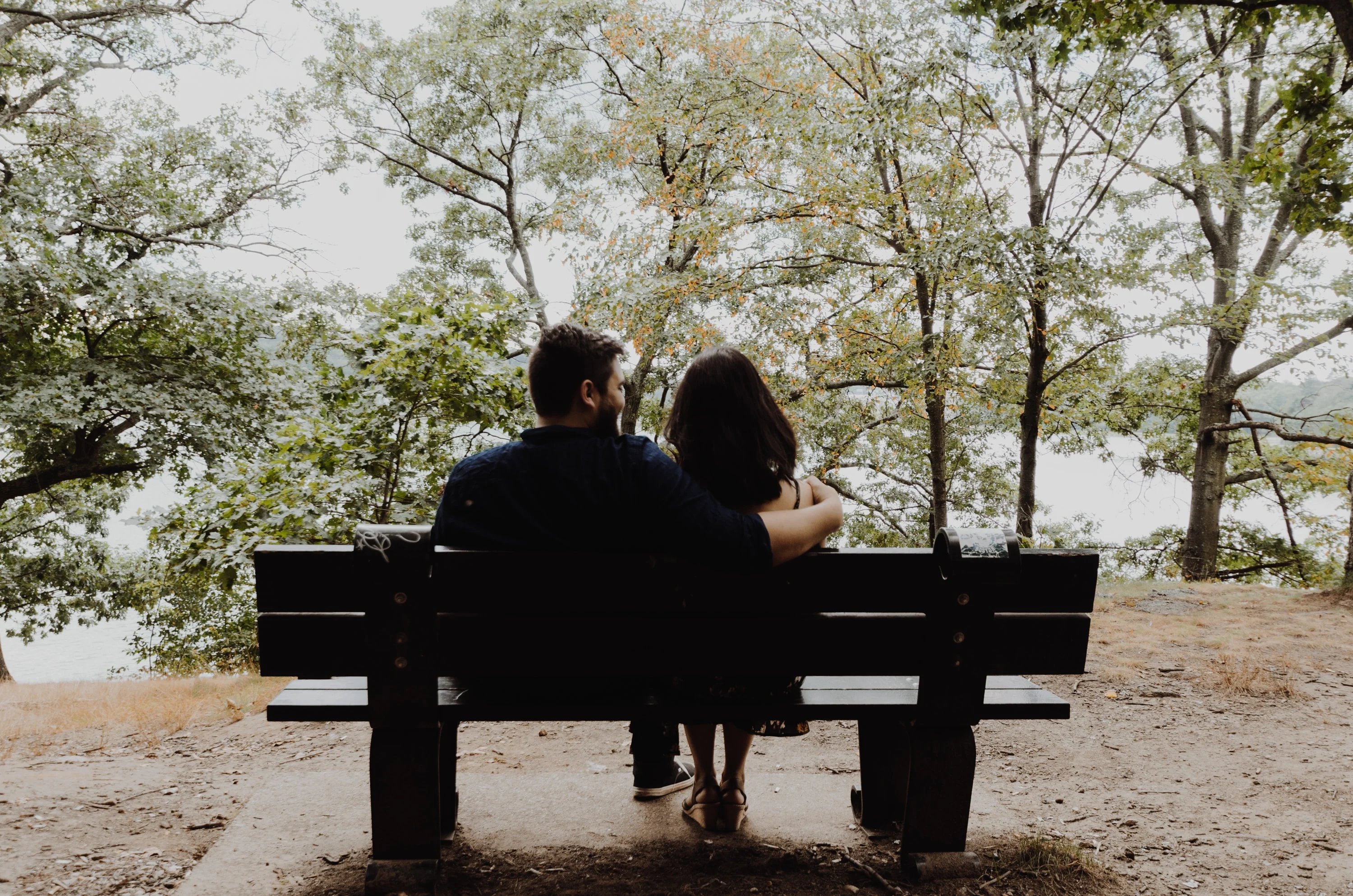 Couple Embracing on a Bench in Nature A couple sitting on a bench, embracing each other, with trees and water in the background.