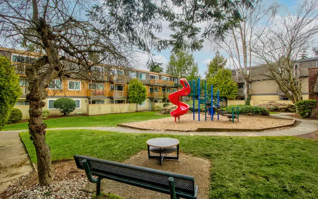 Playground Scene in Residential Area A playground area surrounded by trees and residential buildings. The playground features a red slide and blue climbing structures, with a bench and a circular stone area in the foreground.