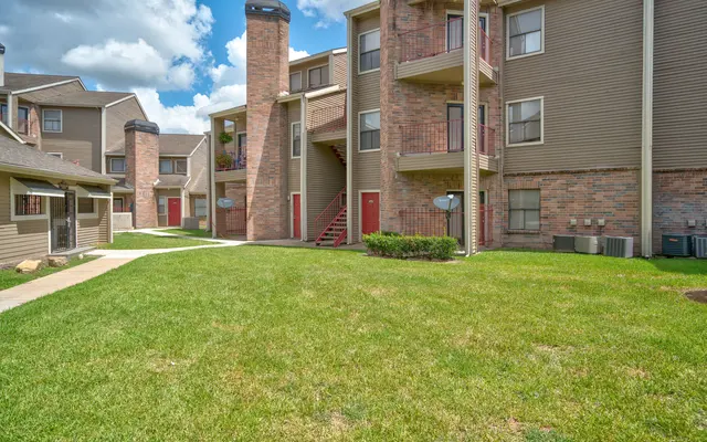 A well-maintained apartment complex courtyard featuring green grass, walkways, and several buildings with a mix of brick and siding exteriors. The scene includes two levels of apartments with balconies, a chimney structure, and partly cloudy skies above.