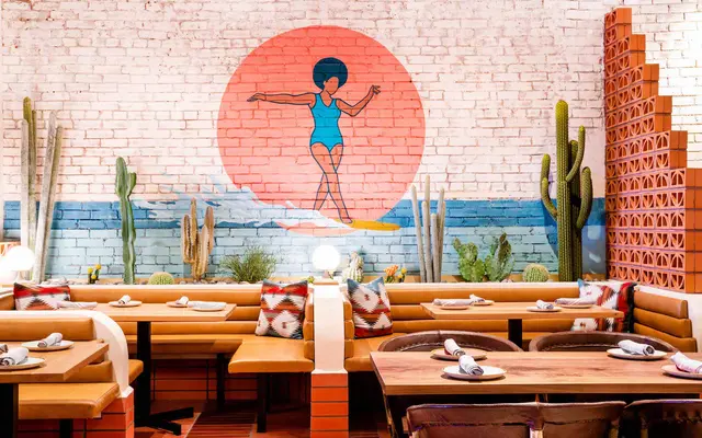 A vibrant restaurant interior featuring a colorful mural of a woman with an afro in front of a large sun, surrounded by cacti and a blue ocean. The seating includes wooden benches and tables set with tableware.