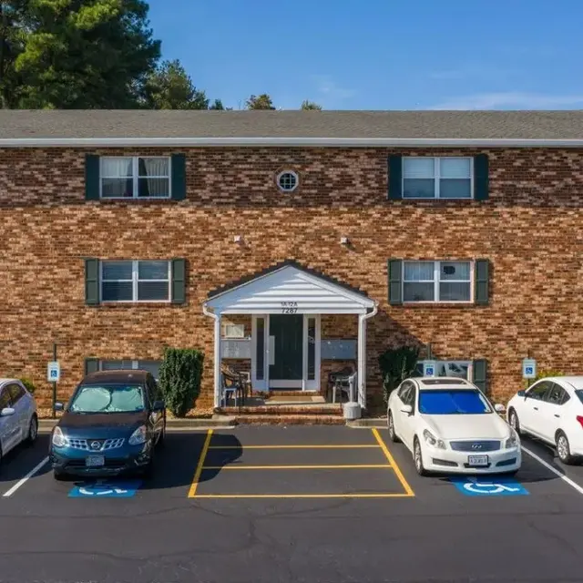 A brick apartment building with multiple windows and a center entrance, surrounded by several parked cars, including two in handicapped spaces.