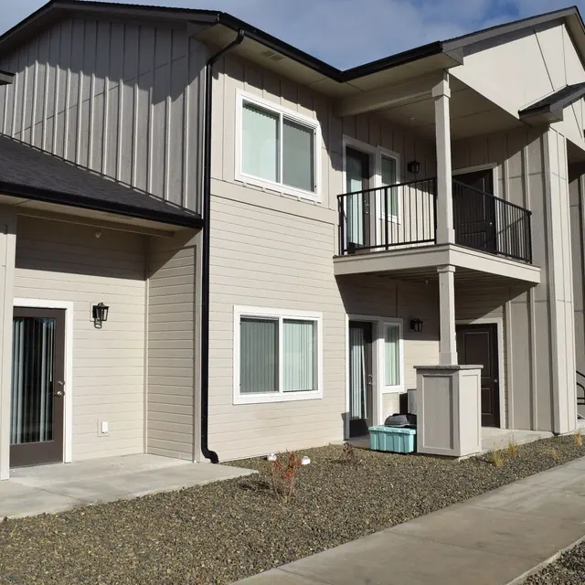 Contemporary Apartment Building A modern two-story apartment building with a combination of light and dark siding, featuring multiple windows, a balcony on the second floor, and a pathway leading up to the entrance.