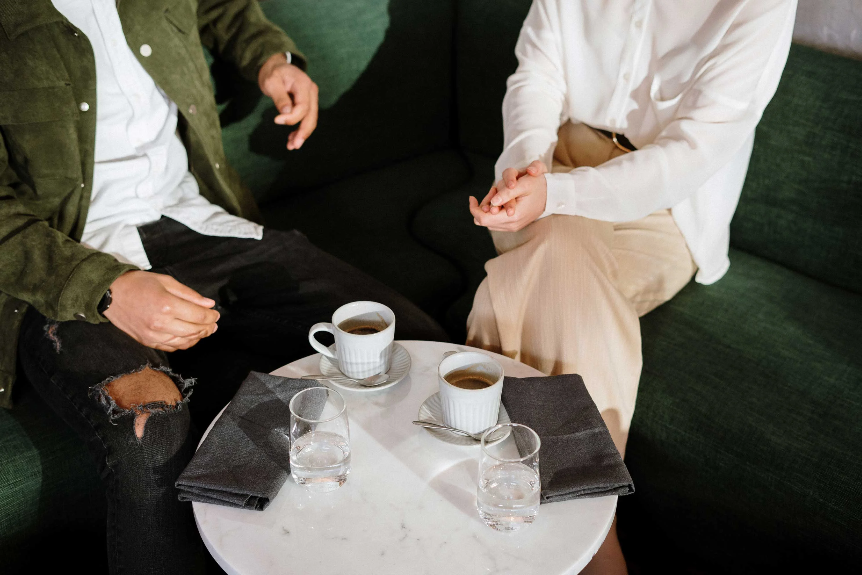 A close-up view of two people seated at a table with coffee cups, water glasses, and napkins. The table is marble, and they are engaged in conversation, with one person gesturing.