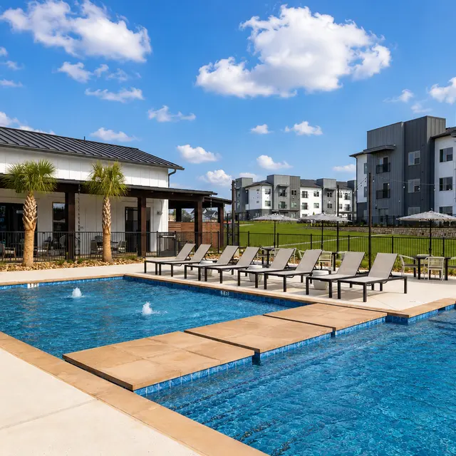 A sunny outdoor residential pool area featuring two swimming pools, lounge chairs, and modern apartments in the background.