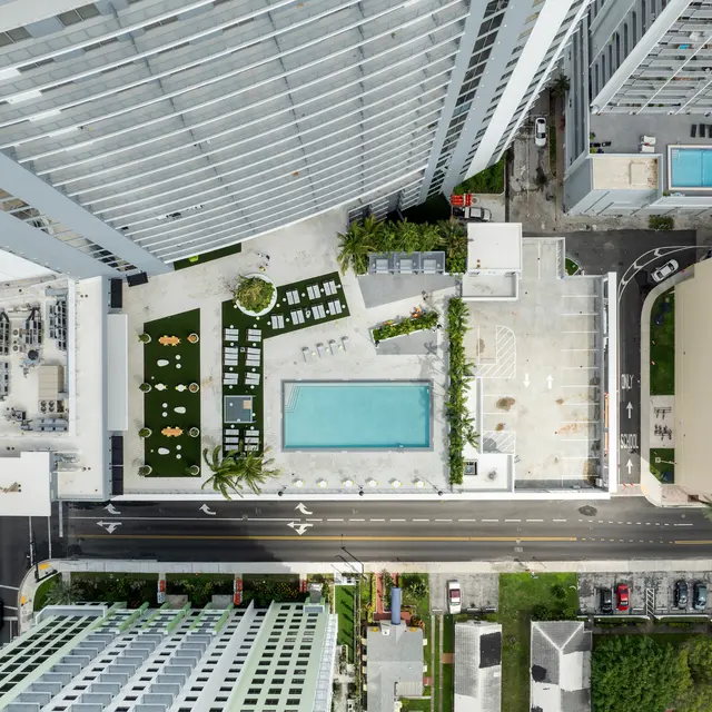 Aerial view of a modern high-rise building overlooking a swimming pool and landscaped area with lounge chairs. Nearby urban streets and another building with a basketball court can be seen.