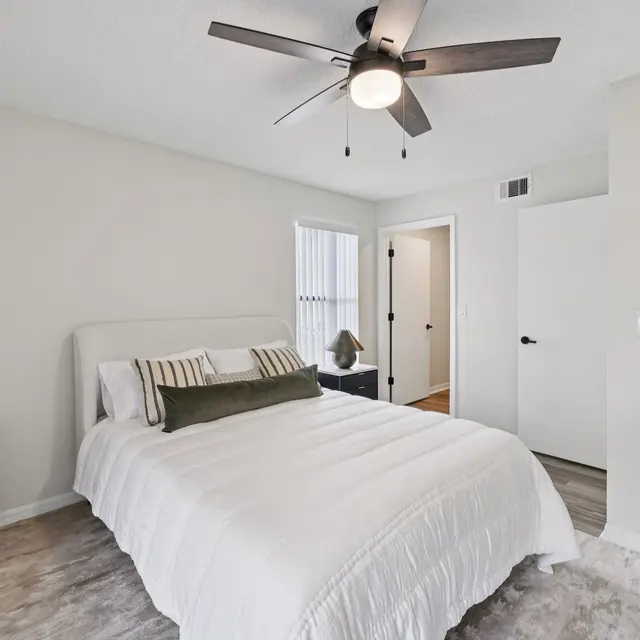 A modern and stylish bedroom featuring a double bed with white bedding, decorative cushions, and a bedside lamp on a dark nightstand. The room has light-colored walls, a ceiling fan, and hardwood flooring with a light area rug.