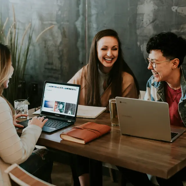 Three people sitting at a wooden table in a cozy cafe, working on laptops and laughing. One person has a laptop open showing a webpage, another is writing in a notebook, and the third is chatting animatedly. There are plants in the background and warm lighting.