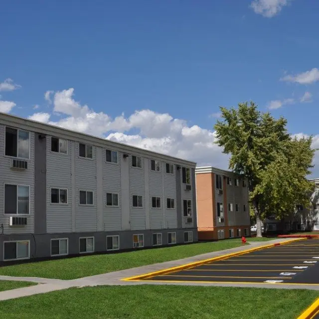 A view of a multi-story apartment complex with grass and trees, under a clear blue sky with some clouds.