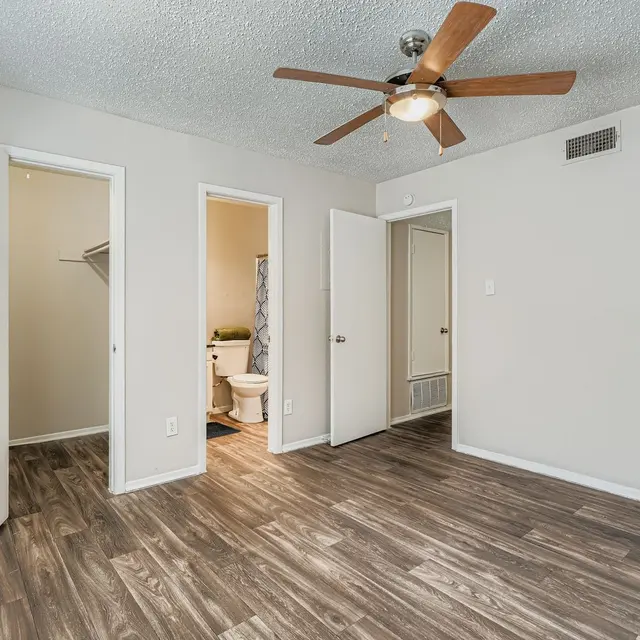 A spacious, well-lit bedroom featuring wooden flooring, a ceiling fan, and two open doors leading to different areas. One door shows a bathroom, while the other leads to a closet.