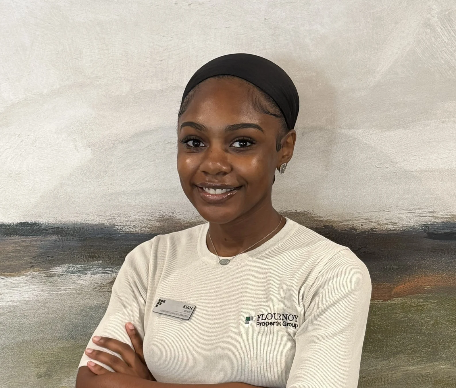 Office Representative at Flournoy Properties A young woman standing with her arms crossed and smiling, wearing a light-colored shirt with a name tag in an office setting.