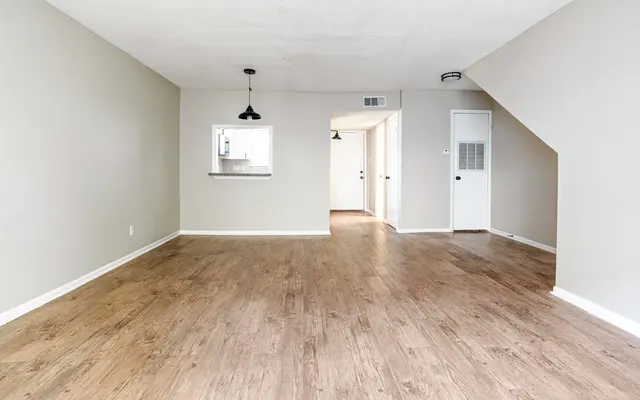 A spacious, empty living room with light gray walls, hardwood flooring, and a pendant light. A doorway leads to another room, and a small window passes through to a kitchen area.
