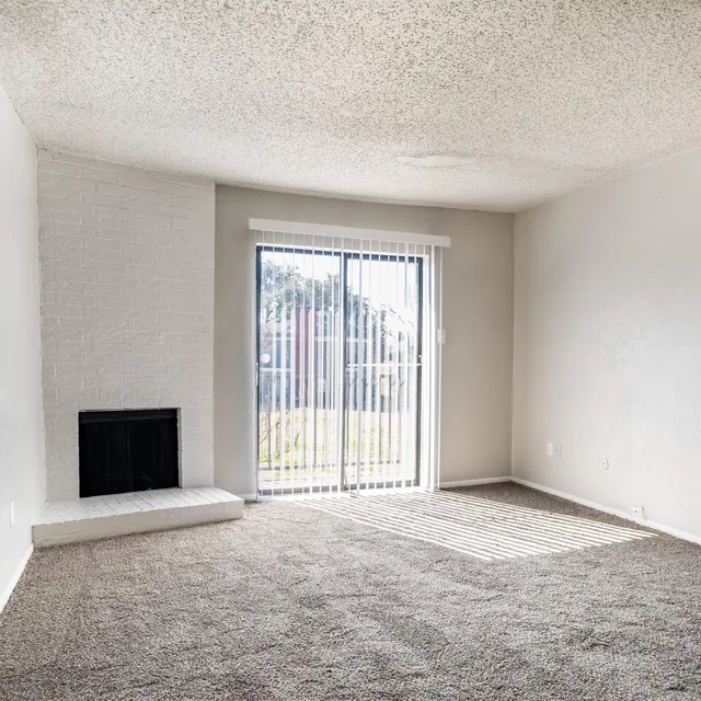 An empty living room with carpeted flooring and a large window with vertical blinds letting in sunlight. There is a small fireplace to the left of the window. The walls are painted white and the ceiling has a textured finish.