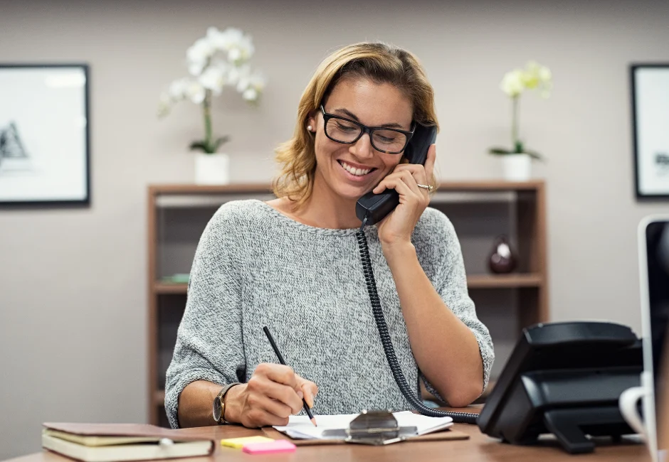 A woman with glasses smiling while sitting at a desk, holding a phone to her ear and taking notes.