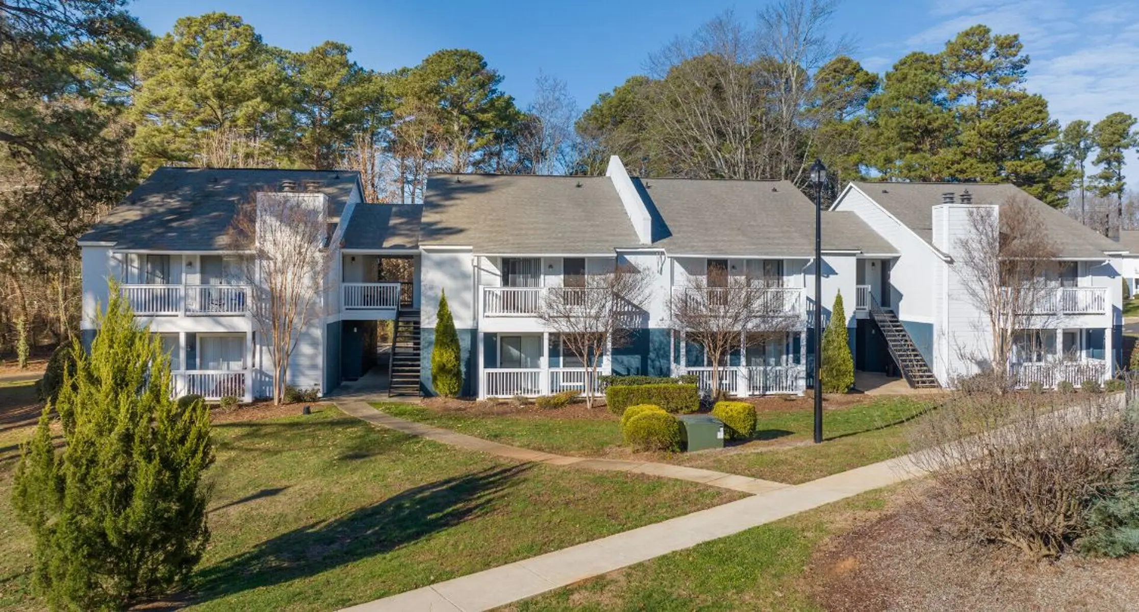 A view of a multi-unit apartment complex surrounded by trees and green landscaping. The buildings have balconies and are arranged in two rows with a pathway leading through the front.