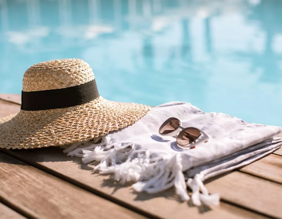 Summer Poolside Accessories A straw sun hat with a black band, a white beach towel with fringes, and a pair of sunglasses placed on a wooden deck beside a swimming pool.