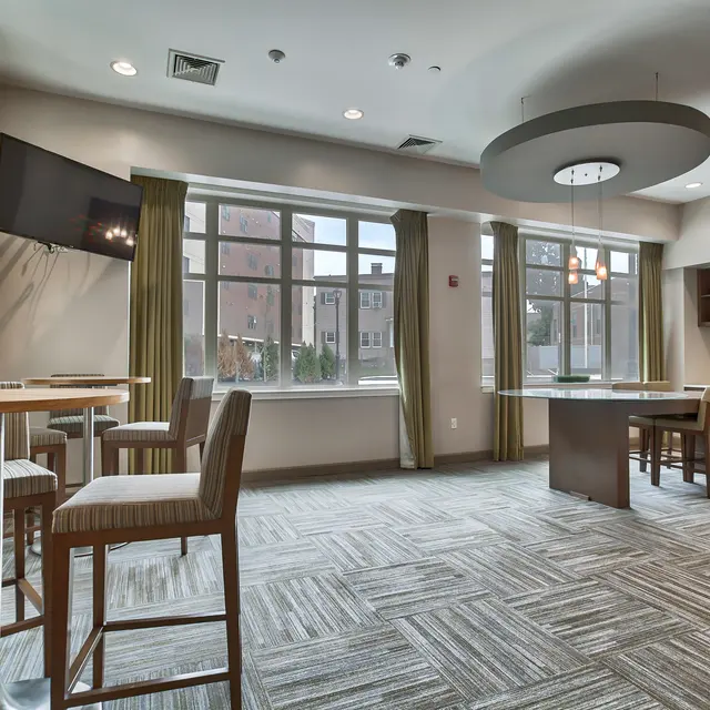 Modern Lounge Area A modern lounge area featuring high-top tables and chairs, large windows with green curtains, and a kitchenette against one wall. A wall-mounted television is visible to one side, and the flooring is a patterned carpet.