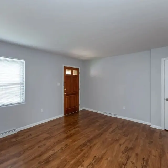 An empty room with light gray walls, wooden flooring, a window with blinds, a brown front door, and a white closet door.