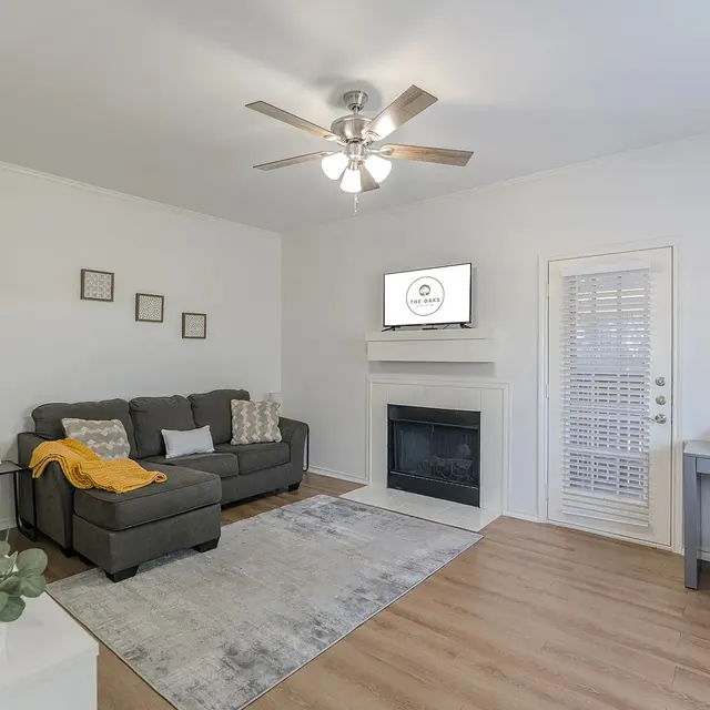 A modern living room featuring a gray sofa with a yellow throw, a fireplace, a TV mounted on the wall, and a desk area with a small plant