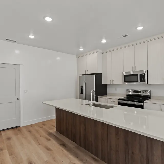 A modern kitchen featuring a large island with a white countertop, dark wooden base, stainless steel appliances including a refrigerator and oven, light wood flooring, and white cabinetry.