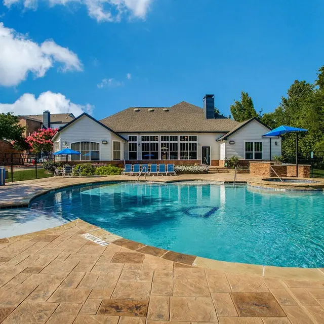 A scenic swimming pool area with a large pool, lounge chairs, and a clubhouse in the background. The pool has a light blue water surface reflecting the sky, surrounded by stone tiles. There are green trees and colorful flowers enhancing the outdoor setting.