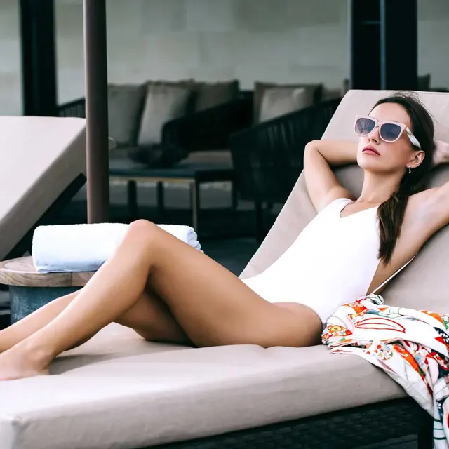 A woman in a white swimsuit lounging on a sunbed, wearing sunglasses, with her arms behind her head and legs stretched out. In a relaxed outdoor setting with plush seating in the background.