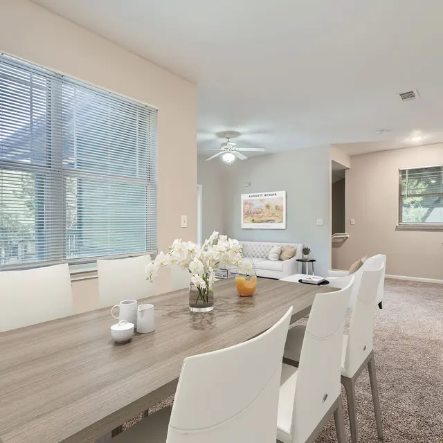 A modern dining area with a wooden table and white chairs, featuring a vase of flowers. In the background, there is a cozy living room with a sofa, a ceiling fan, and a window with blinds.