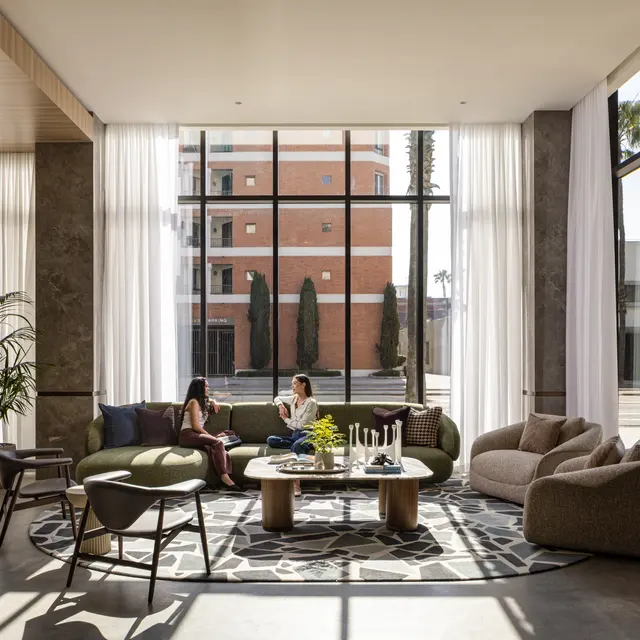 A modern lobby with large windows allowing natural light, featuring a circular rug, two women chatting on a green sofa, and various stylish furniture including chairs and tables.