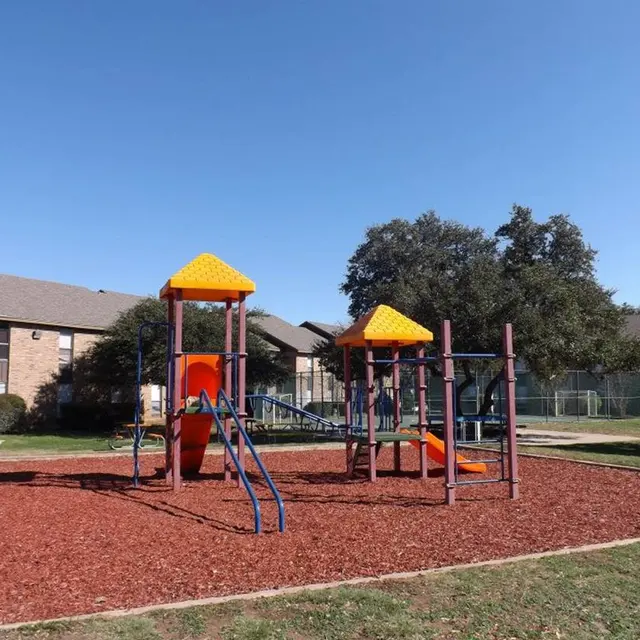 A colorful playground featuring two play structures with yellow roofs, surrounded by red mulch and grassy areas. Nearby are residential buildings and trees.