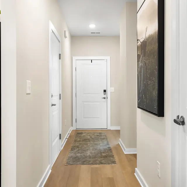 A modern hallway in a home featuring light walls, a large area rug, and a stainless steel refrigerator on the left side. The entrance door is visible at the end of the hallway, and there are several doors on the left side.