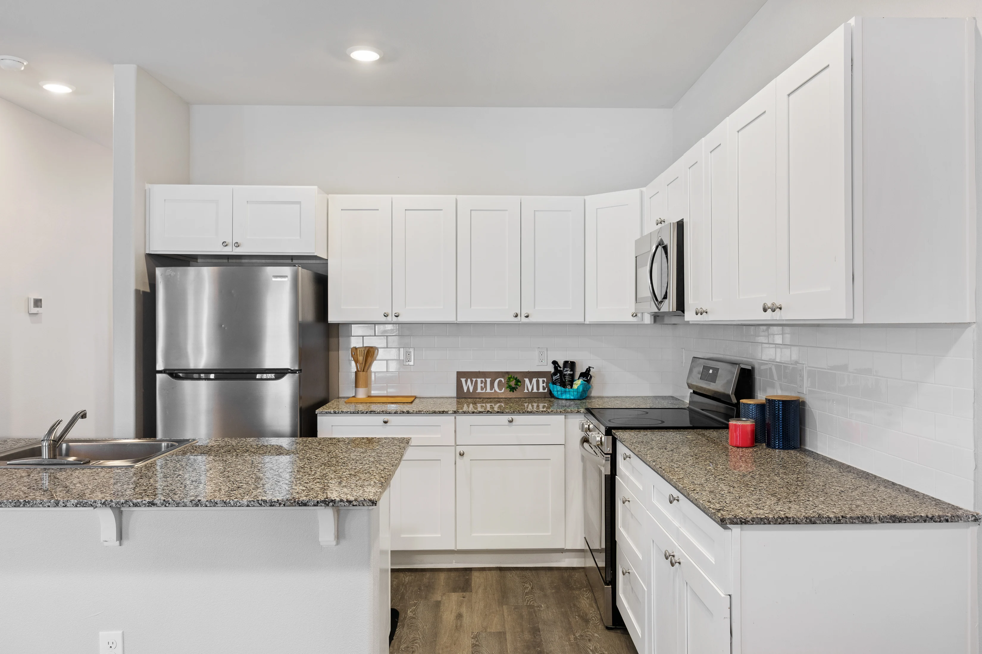 A modern kitchen featuring white cabinetry, stainless steel appliances, and a granite countertop. A sign that reads 'WELCOME' is displayed on the counter along with kitchen utensils.