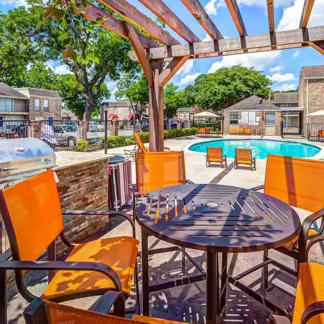 A sunlit poolside area with a round wooden table and orange chairs under a wooden pergola. In the background, there's a swimming pool and several lounge chairs.