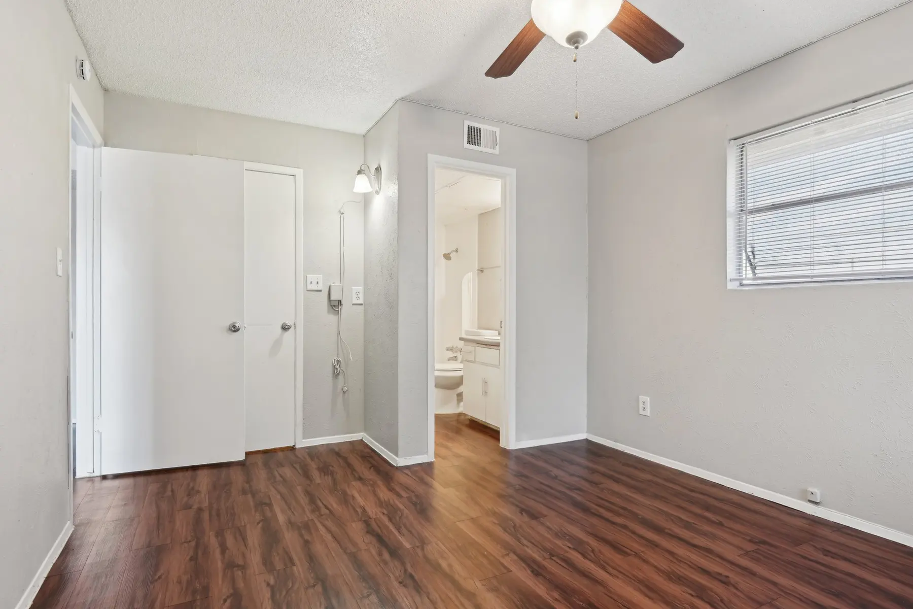 A small empty room with wood flooring, white walls, and a ceiling fan. An open doorway leads to a bathroom area.